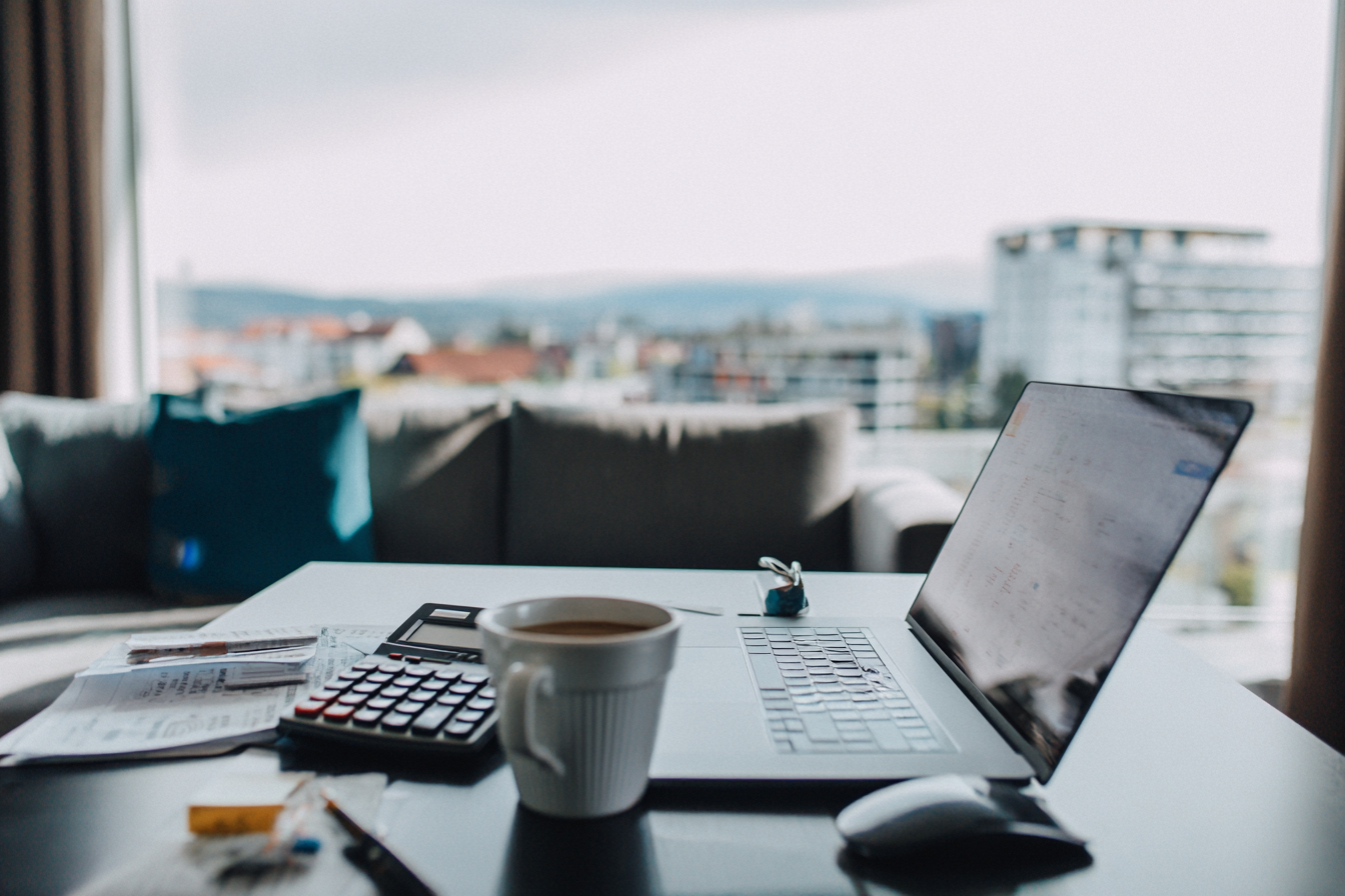 Ordinateur portable, calculatrice, tasse de café et justificatifs sur une table devant une fenêtre donnant sur la ville