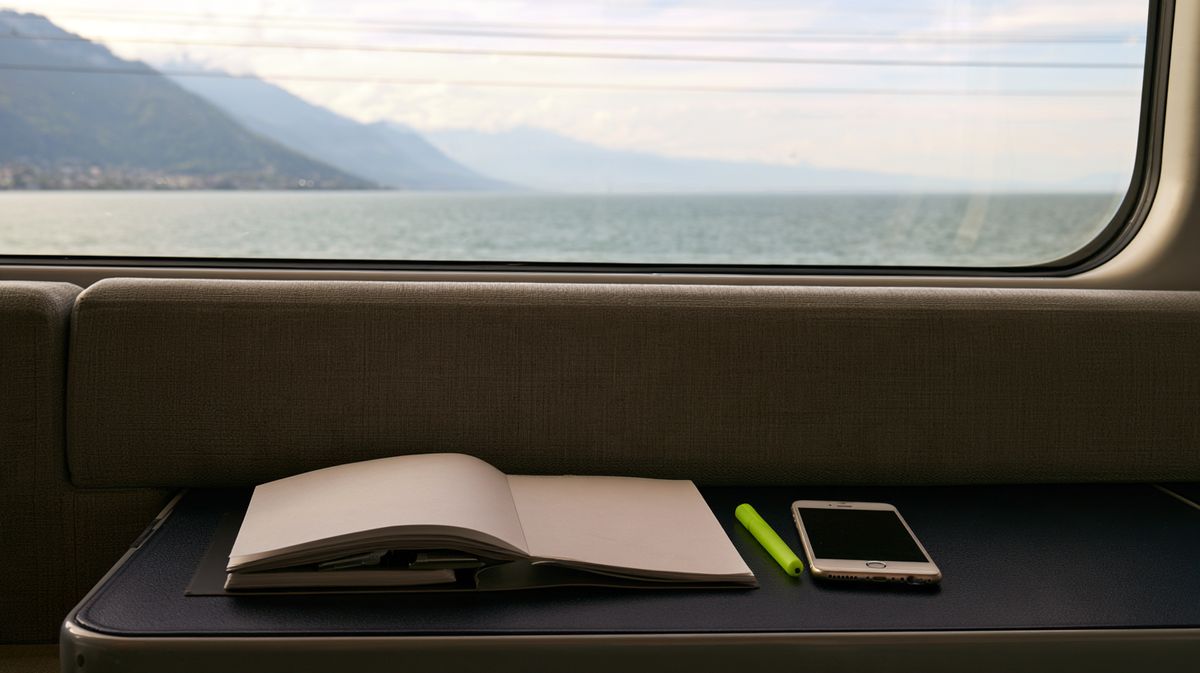 Blick aus dem Zugfenster auf einen See und Berge im Dunst, auf dem Tischchen ein offenes Notizbuch, ein Stift und ein Handy