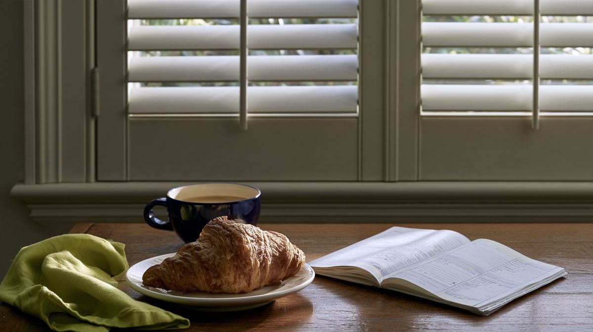 Croissant, tasse de café et cahier ouvert sur une table en bois devant des volets fermés, un petit-déjeuner en Suisse romande