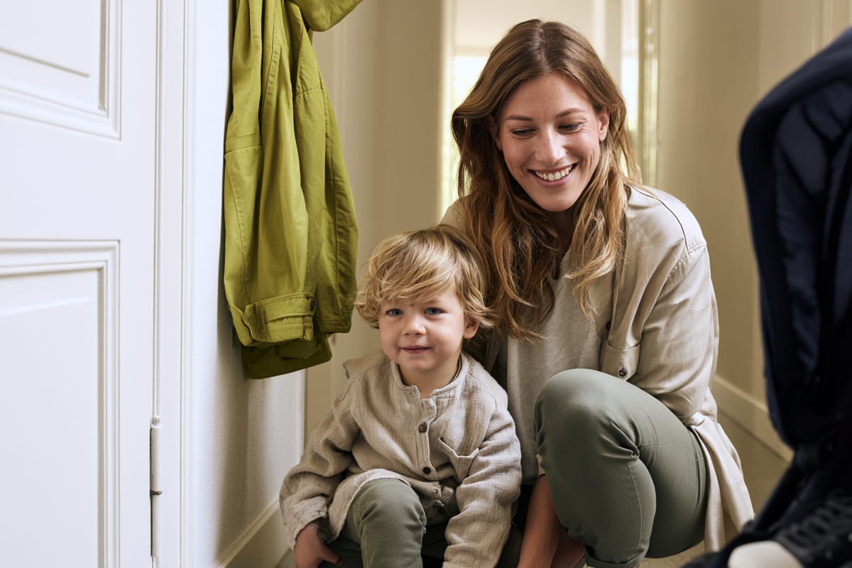 A nanny crouching in the hallway next to a toddler, ready to go out, a jacket on the hook and a pushchair in the foreground