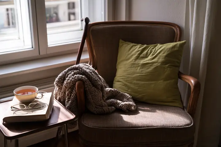 Sessel am Fenster einer älteren Wohnung mit Strickdecke, Lesebrille, Buch, Tee und Gehstock